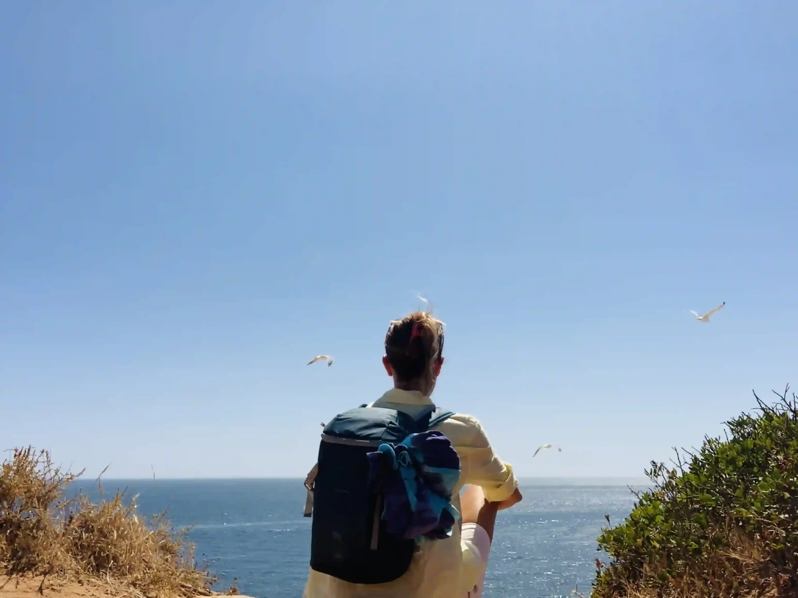 Persona di viaggiinchiaro con zaino in spalla che osserva l’oceano dalla scogliera dell’Algarve, in Portogallo, con gabbiani in volo e cielo azzurro